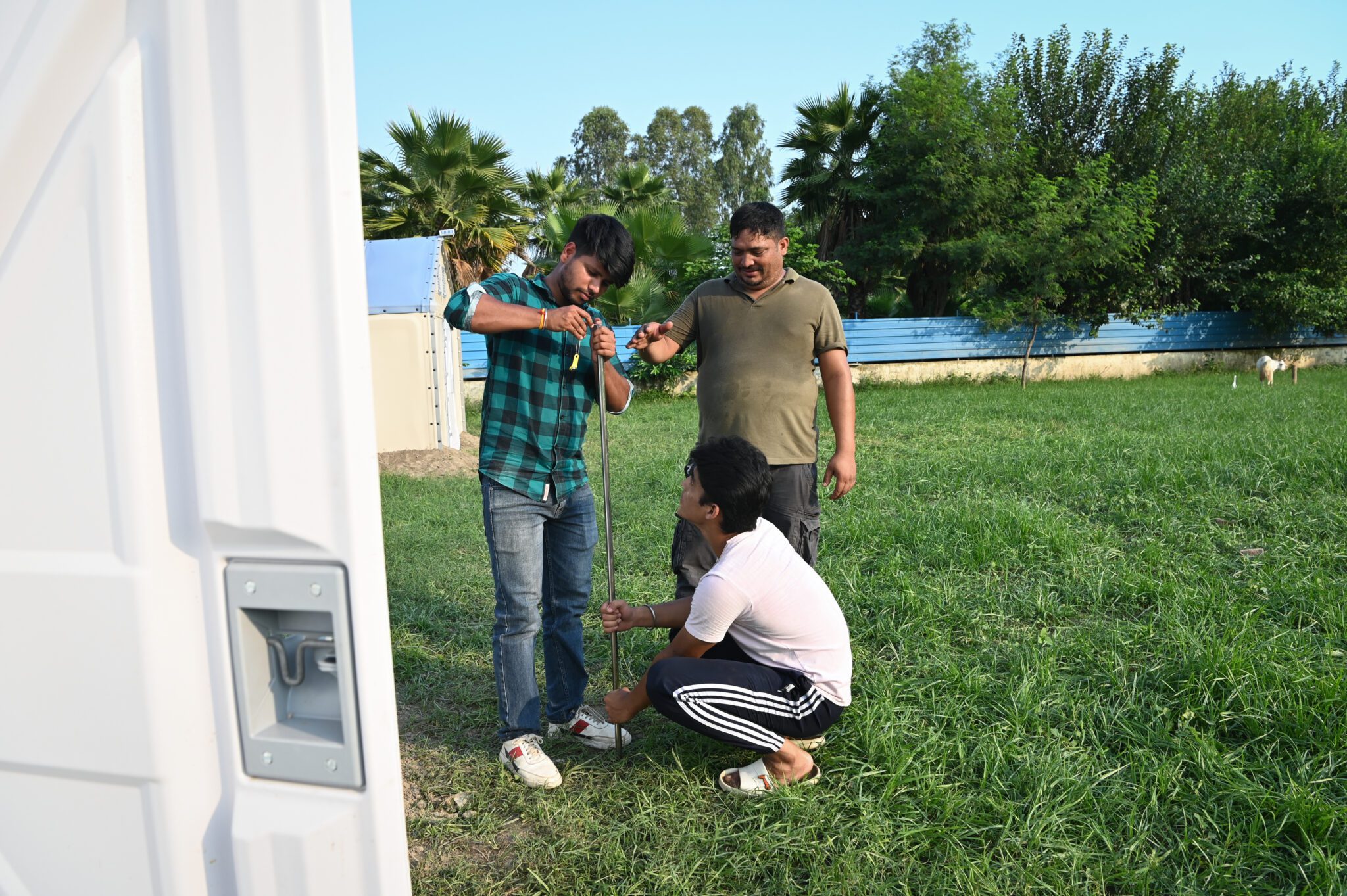 Every degree matters: testing and improving shelter temperatures with partners in India Three men setting up a metal pole on the grass beside a shelter.